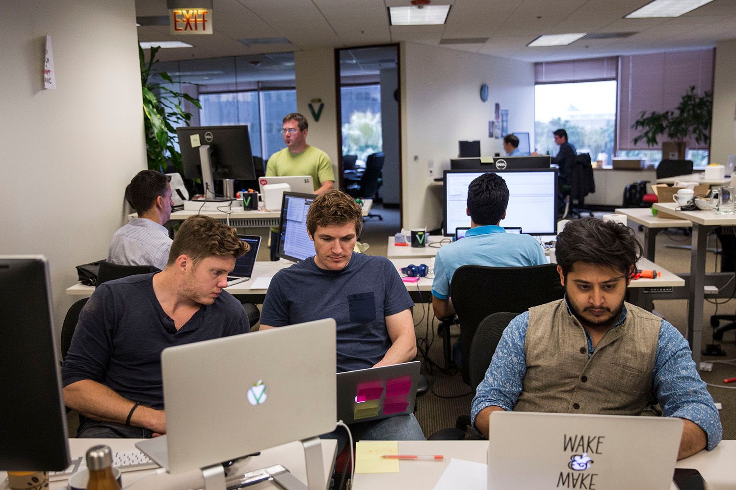 Adult men in office setting, open floor plan, apple laptop computers.