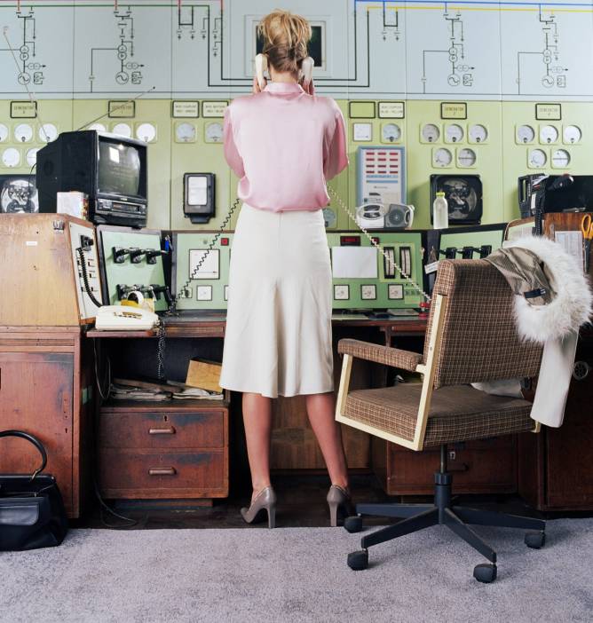 Woman standing at a desk holding two corded phones with her back facing the camera, indicating multitasking