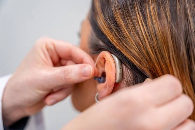 Doctor placing a small hearing aid to the ear of a young woman.