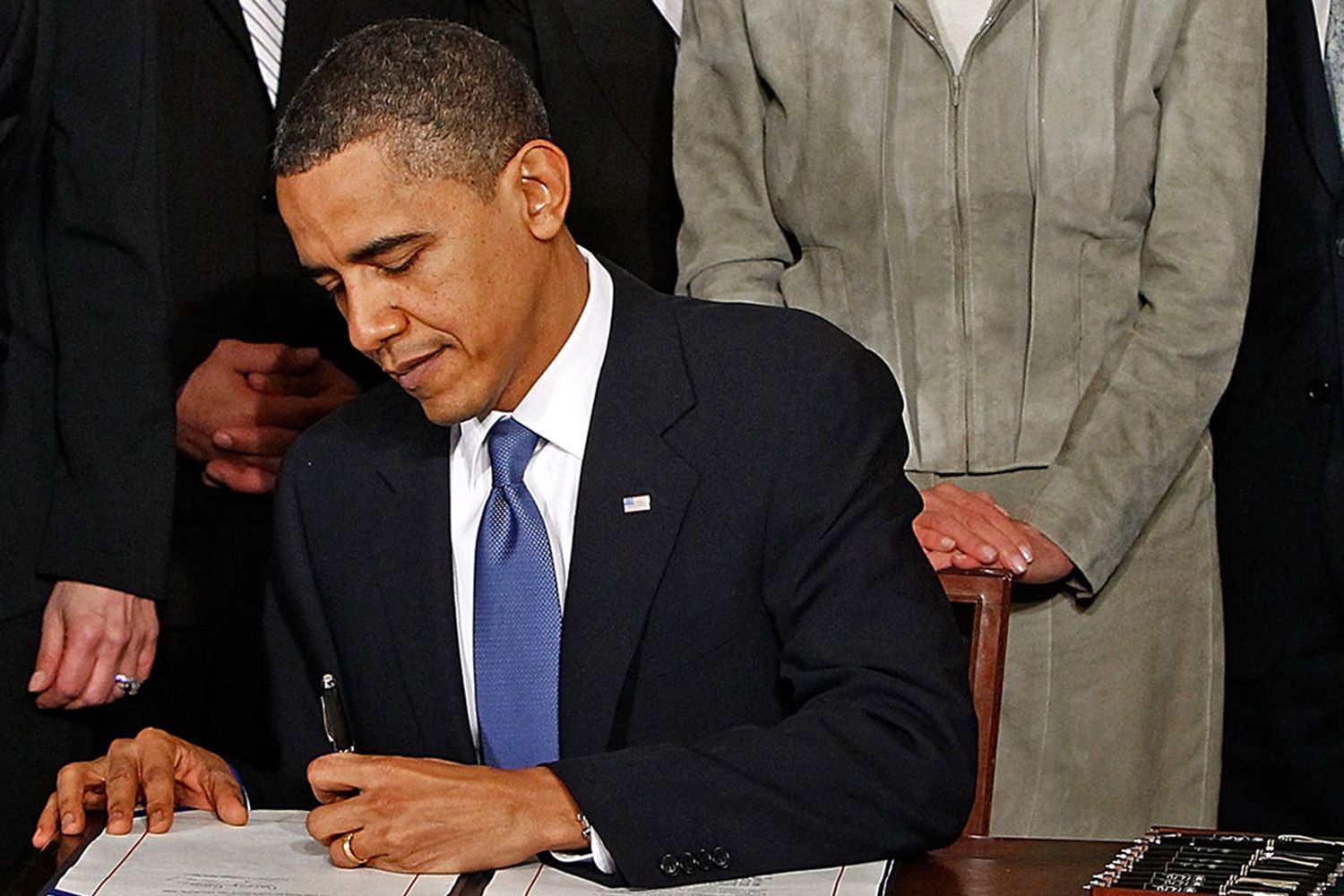 President Barack Obama signs an official document, people stand behind him as witnesses.