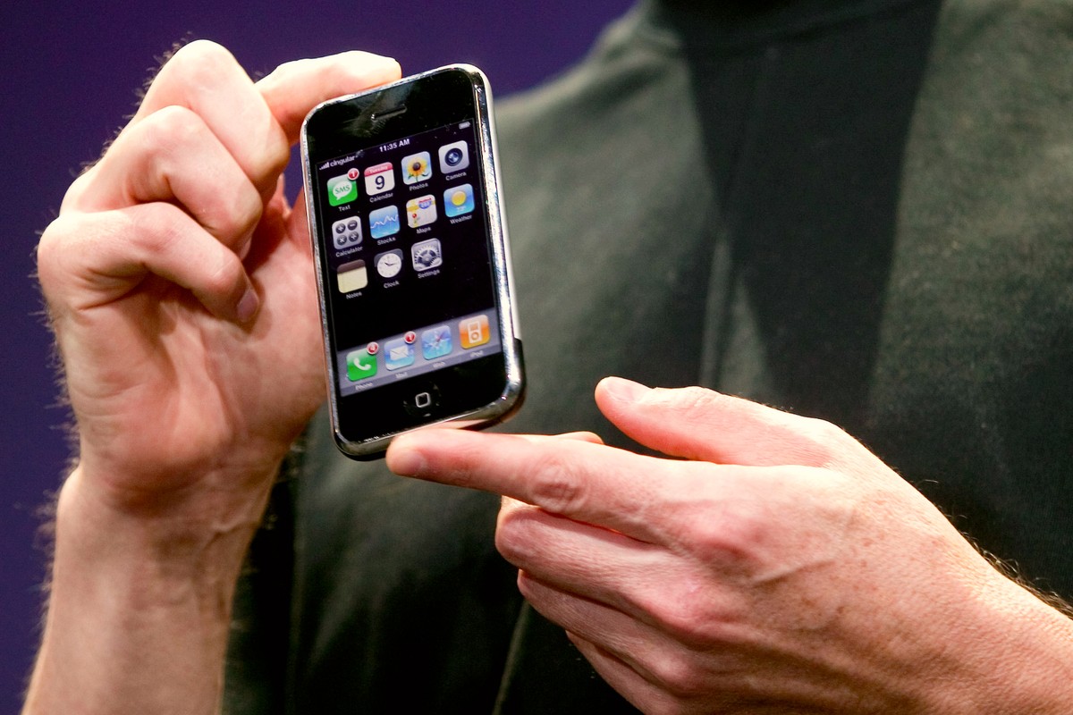 A hand holds the first iteration of the iPhone, a chunky black electronic with a digital screen