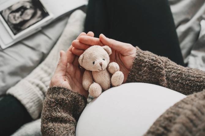 A pregnant person holds a teddy bear on their bump.