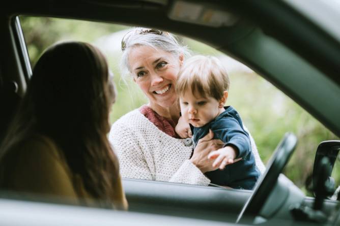 A woman sits in a car while speaking to an older woman holding a toddler.