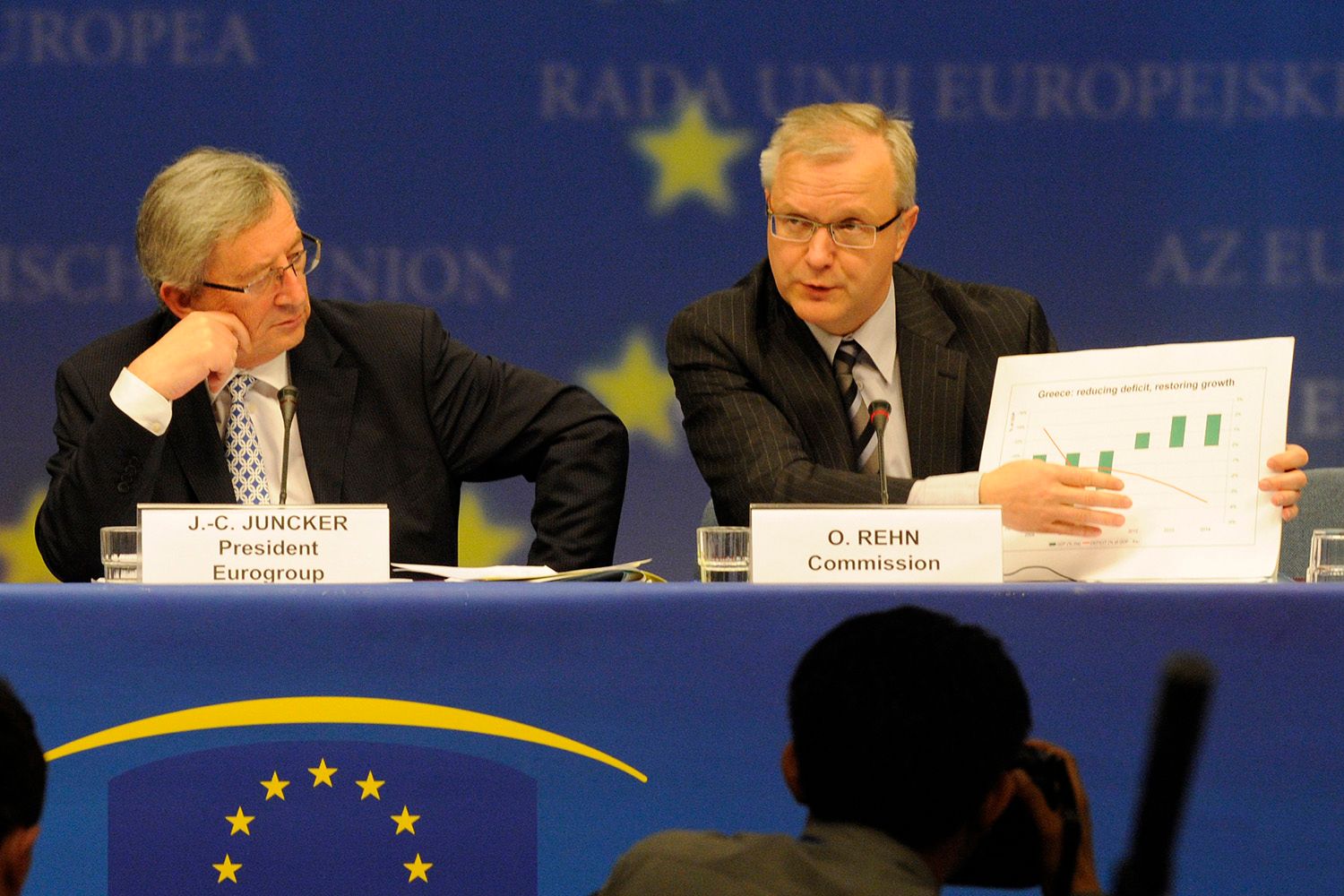 Men in suits presenting to an audience, one man speaks, holding a printed graph, the other listens intently.