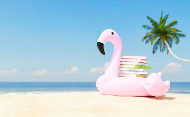 A stack of books are balanced on an inflatable pool float shaped like a flamingo, which sits on a sunny beach.