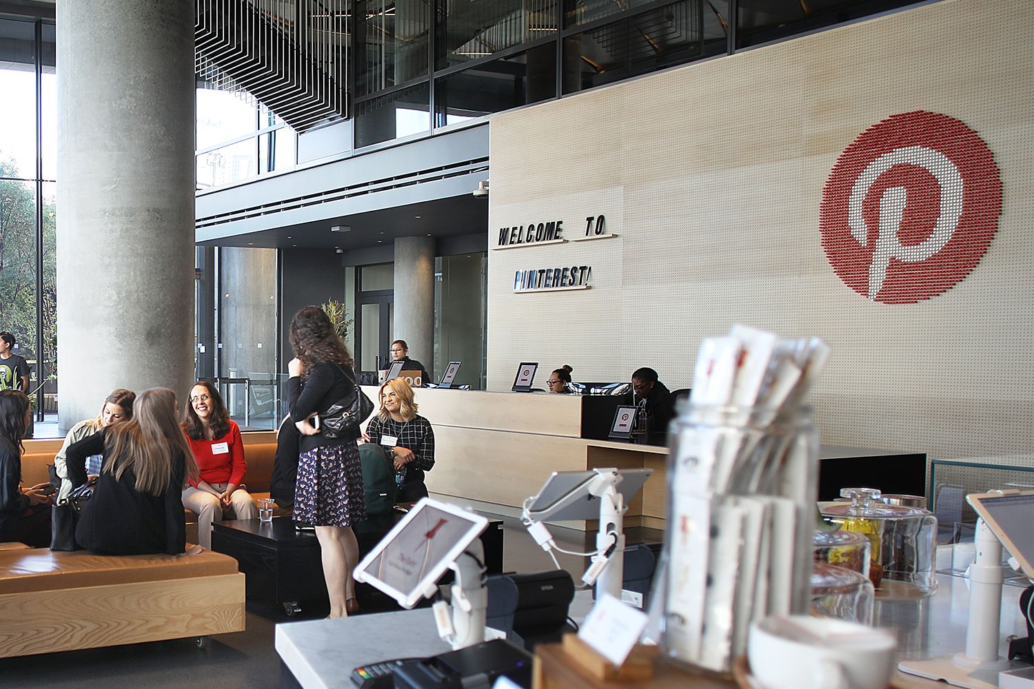 People sitting, talking, mingling in the coffee shop lobby of Pinterest. The logo, a large letter P on a red circle hangs on a wood wall behind the receptionist desk