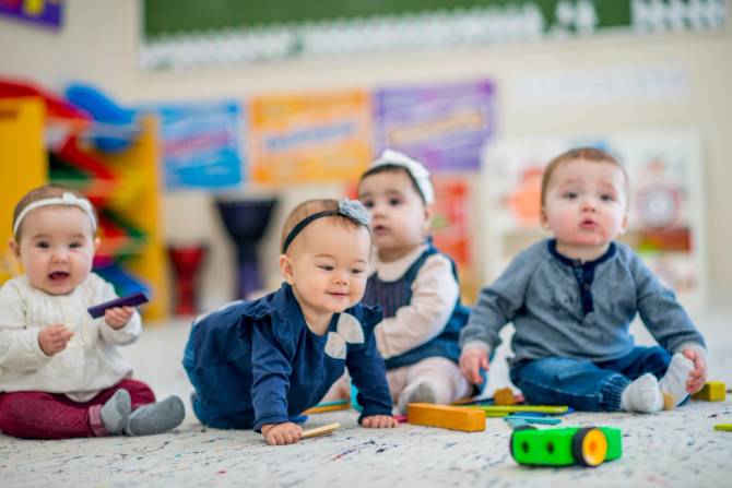 four babies, around six months old in a daycare setting. three have bows on their heads, all looking content