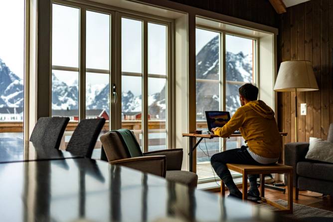 A man works at desk looking out over mountain landscape.
