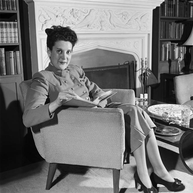 A black and white photo of Sylvia Porter sitting on an accent chair with a book. 