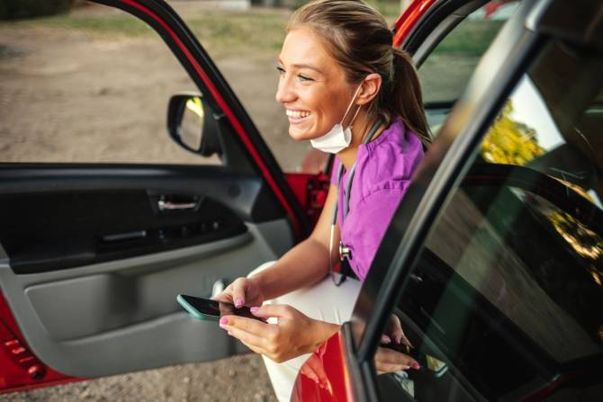 A female-presenting nurse smiles as she steps out of her car, holding an iPhone.