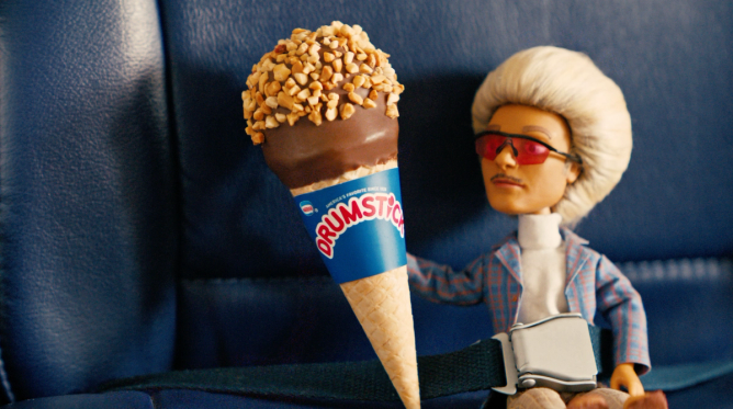 an image of Drumstick ice cream doll mascot Dr. Umstick holding a Drumstick ice-cream cone on a plane seat, in a still from Drumstick's 2024 Super Bowl ad