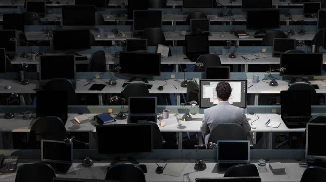 a large, dimly lit office with all empty desks except one with a male worker sitting in his chair looking at his computer screen