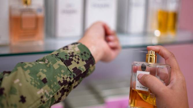 A woman wearing a camo uniform sprays a perfume sample on her wrist. 