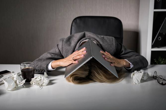 A person face down on their desk hiding under their laptop computer.
