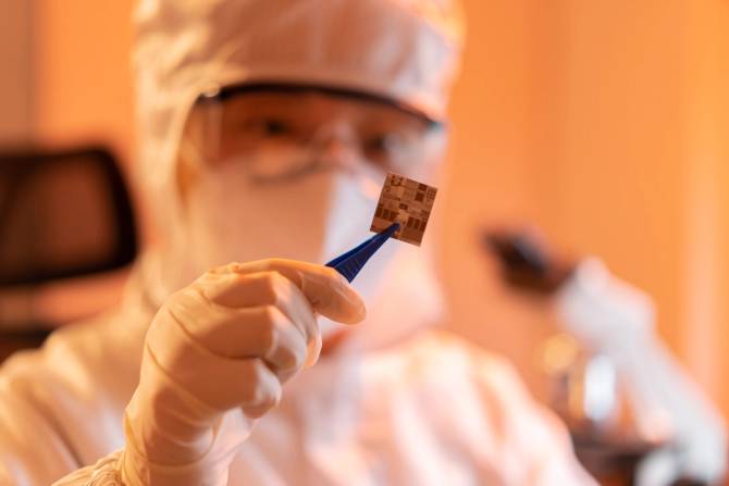 A tech worker in a dust-free lab holds up a microchip with tweezers.
