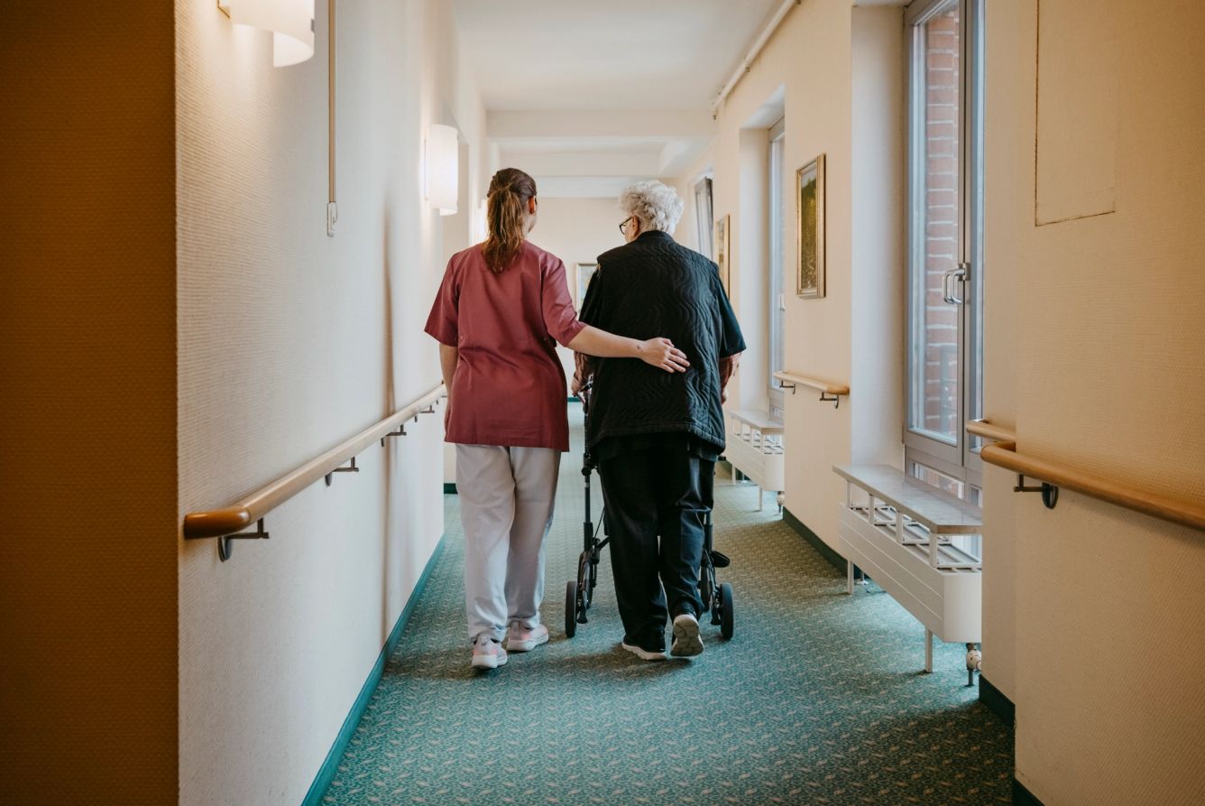 Rear view of female caregiver assisting senior woman walking with mobility walker in corridor