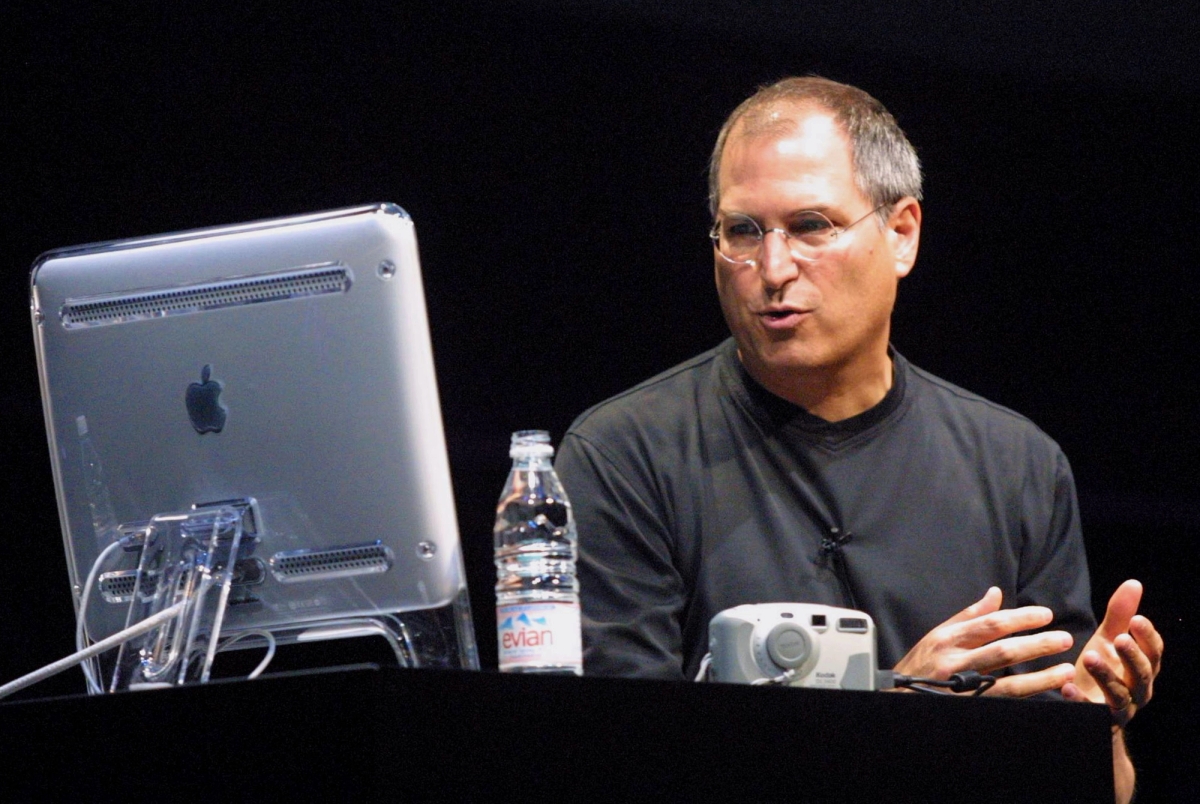 Steve Jobs, in a black t-shirt and delicate wire glasses, speaks while seated in front of a desktop computer.