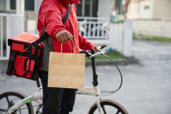 A food delivery person hands a brown paper bag delivery toward the camera, with a bike and an insulated bag visible in the background