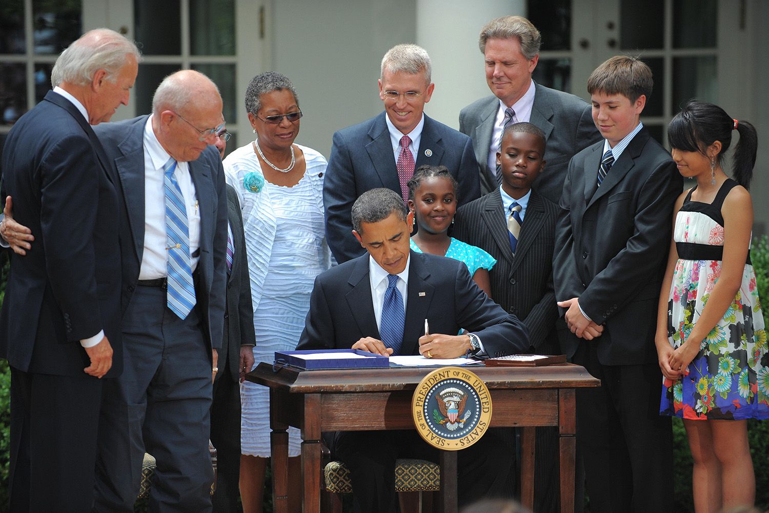Obama signing a bill surrounded by citizens and family members of various ages, races, and genders.