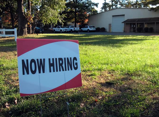 A sign saying "Now Hiring" on a patch of grass in front of a building.