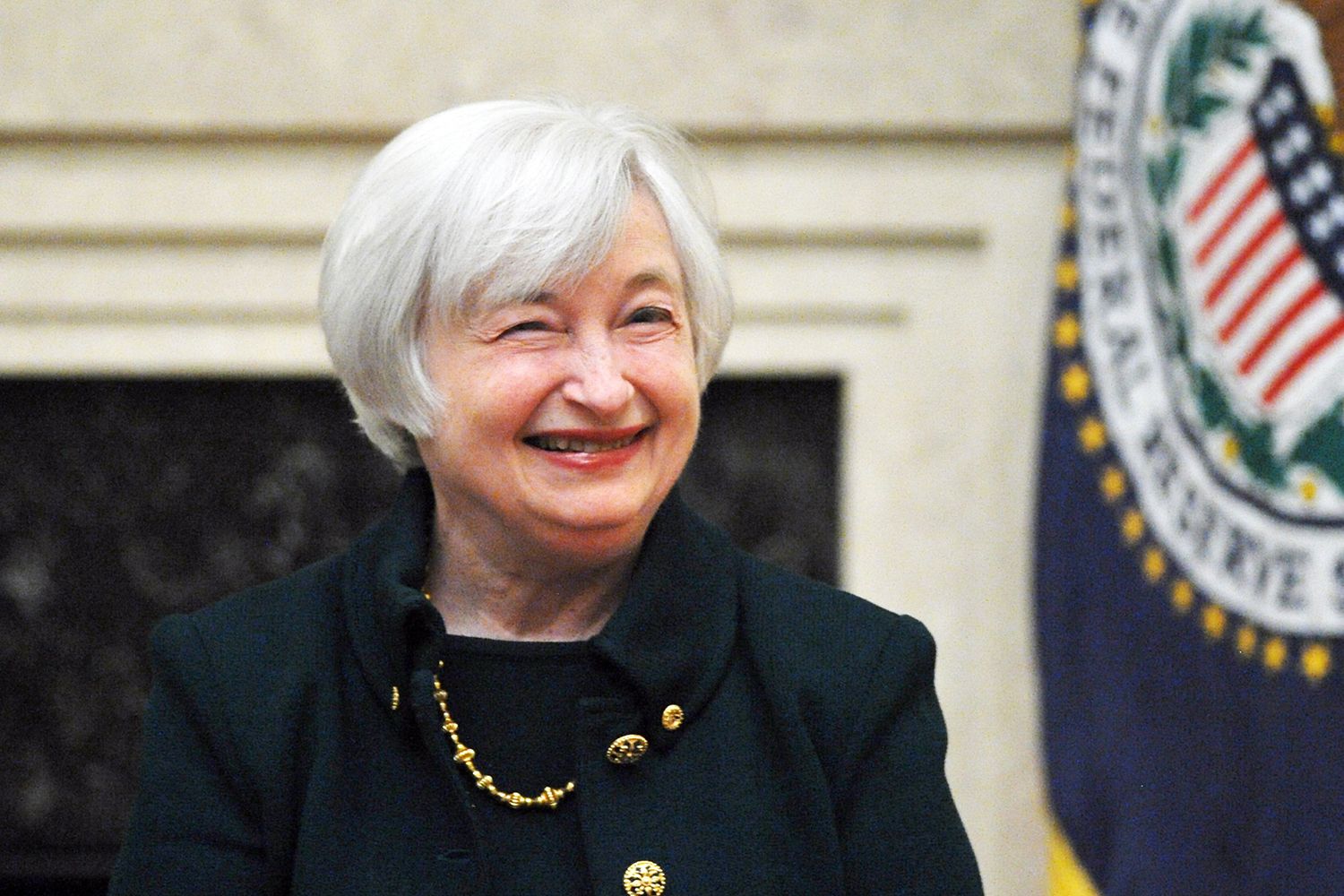 A close-up portrait shot of Janet Yellen, an older woman with short silver hair smiling in front of the Federal Reserve flag