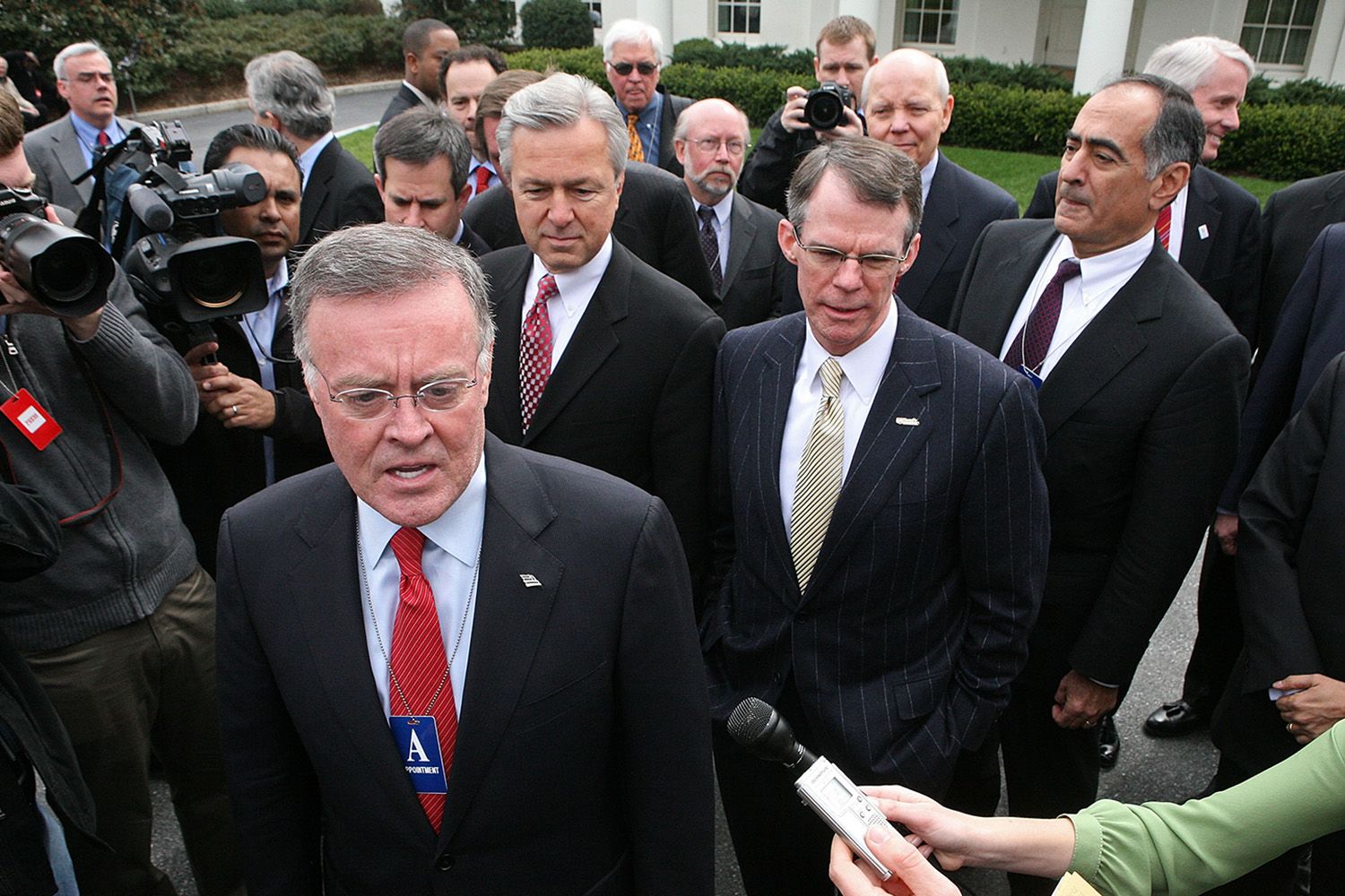 Man in suit with stern expression, press interview, surrounded by men in suits, outdoor procession.