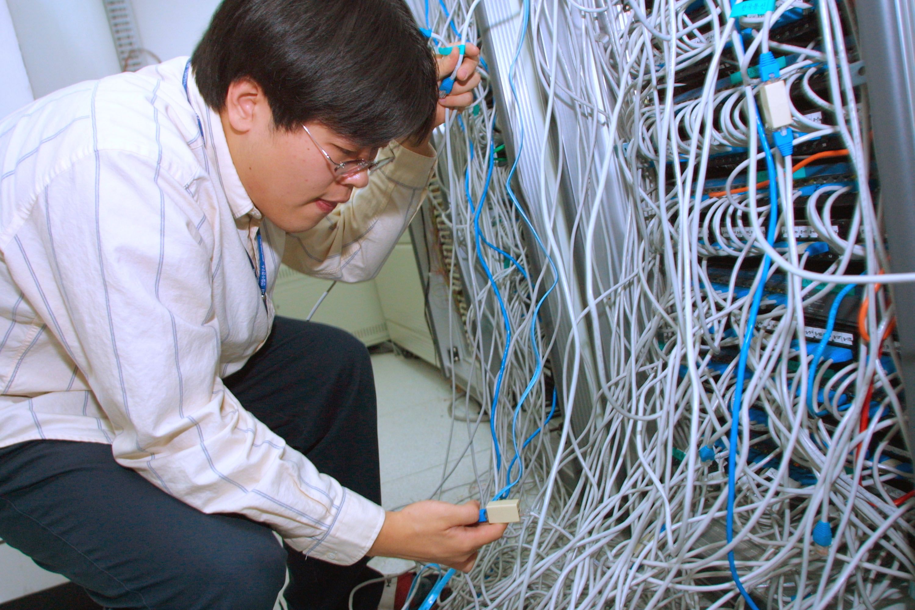 A young professional man crouches down, he appears to be studying a large interwoven set of cables coming from the back of a computer or server.