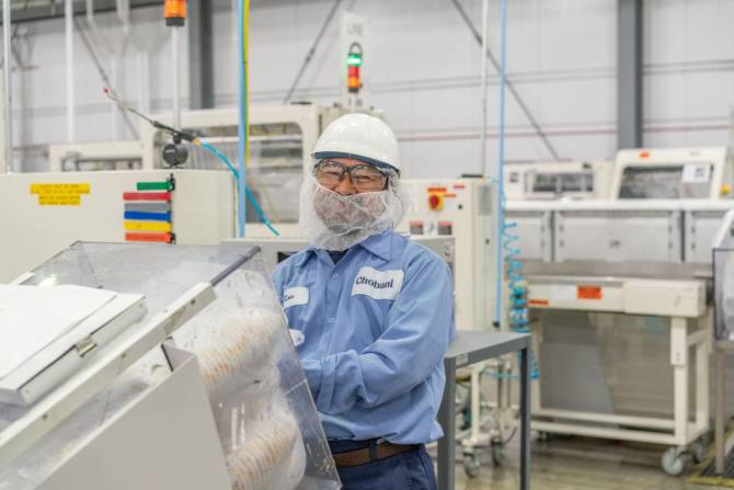 man wearing a light blue Chobani shirt, hard hat and classes smiles smiles as he works at a pice of white machinery in a factory