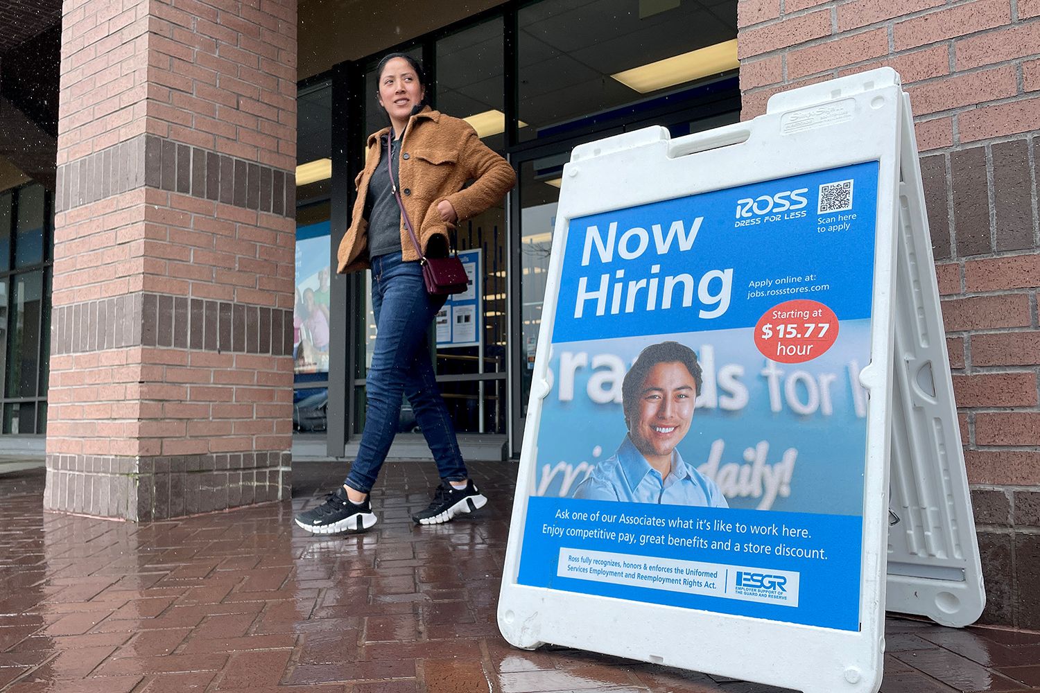 A woman walks out of a building, an A-frame standing board is on the sidewalk with Now Hiring and a hourly rate advertised.