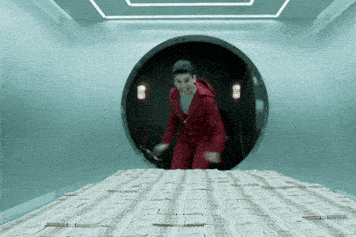 A man in a red jump suit jumping onto a massive pile of money inside a vault