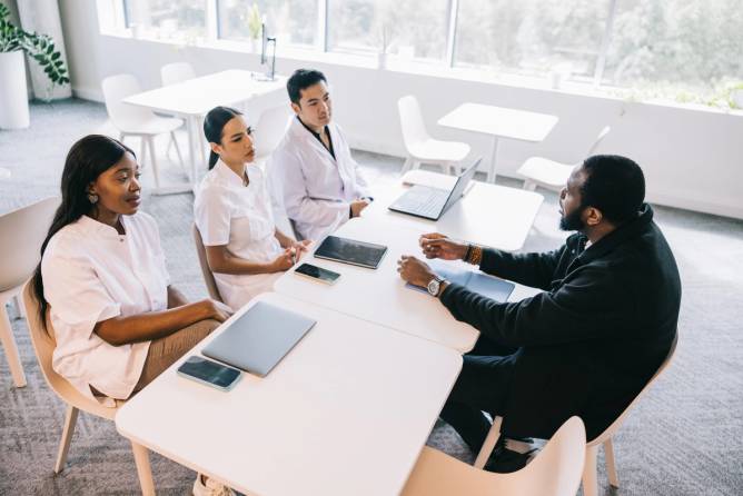 In a hospital conference room, a medical team listens attentively as a new hire discusses their qualifications