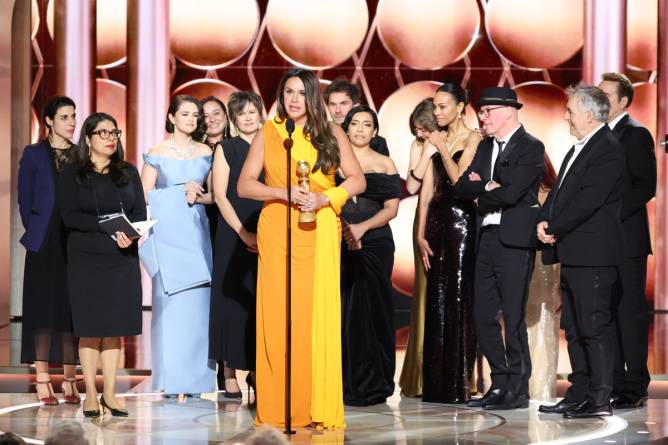 Karla Sofia Gascón holds a Golden Globe award onstage with the rest of the 'Emilia Pérez' cast and crew behind her.