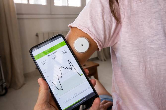 A person holds a phone with glucose monitoring while a person stands in front of them with the patch on, tracking their vitals.