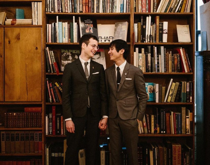 two men holding hands, wearing suits and looking at each other in front of a bookshelf