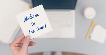 A person’s hand holding a Post It note saying “Welcome to the team!” above a desk with a laptop and office supplies