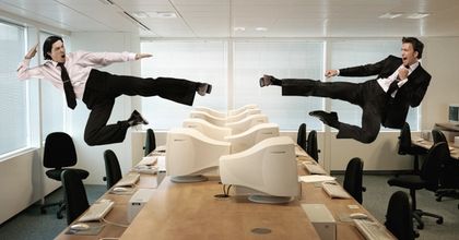 Two businesspeople in an office cubicle performing flying jump kicks at each other over a desk of computer monitors