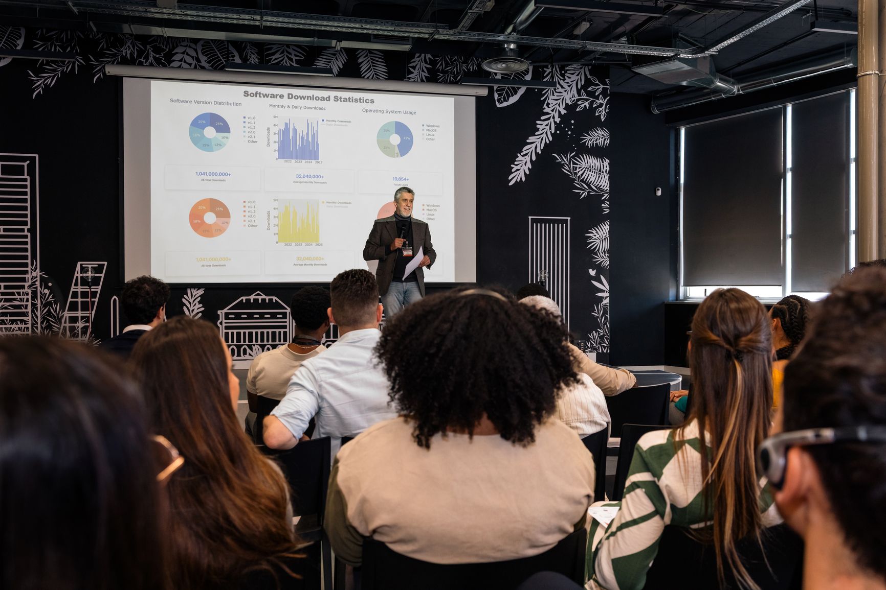 A speaker presents data charts and graphs on a projected screen to a seated audience in a dimly lit modern conference room.