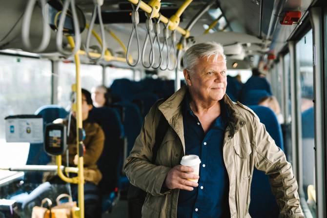 An older man smiles while holding a cup of coffee on a bus.