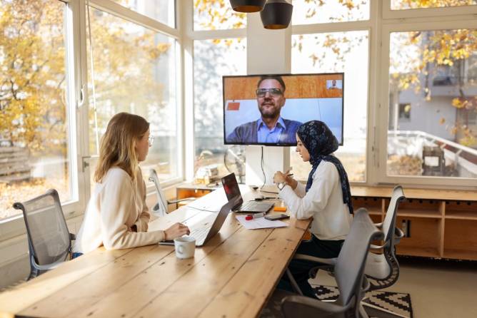 Two workers sit at a table with their laptops while another colleague appears on a video screen.