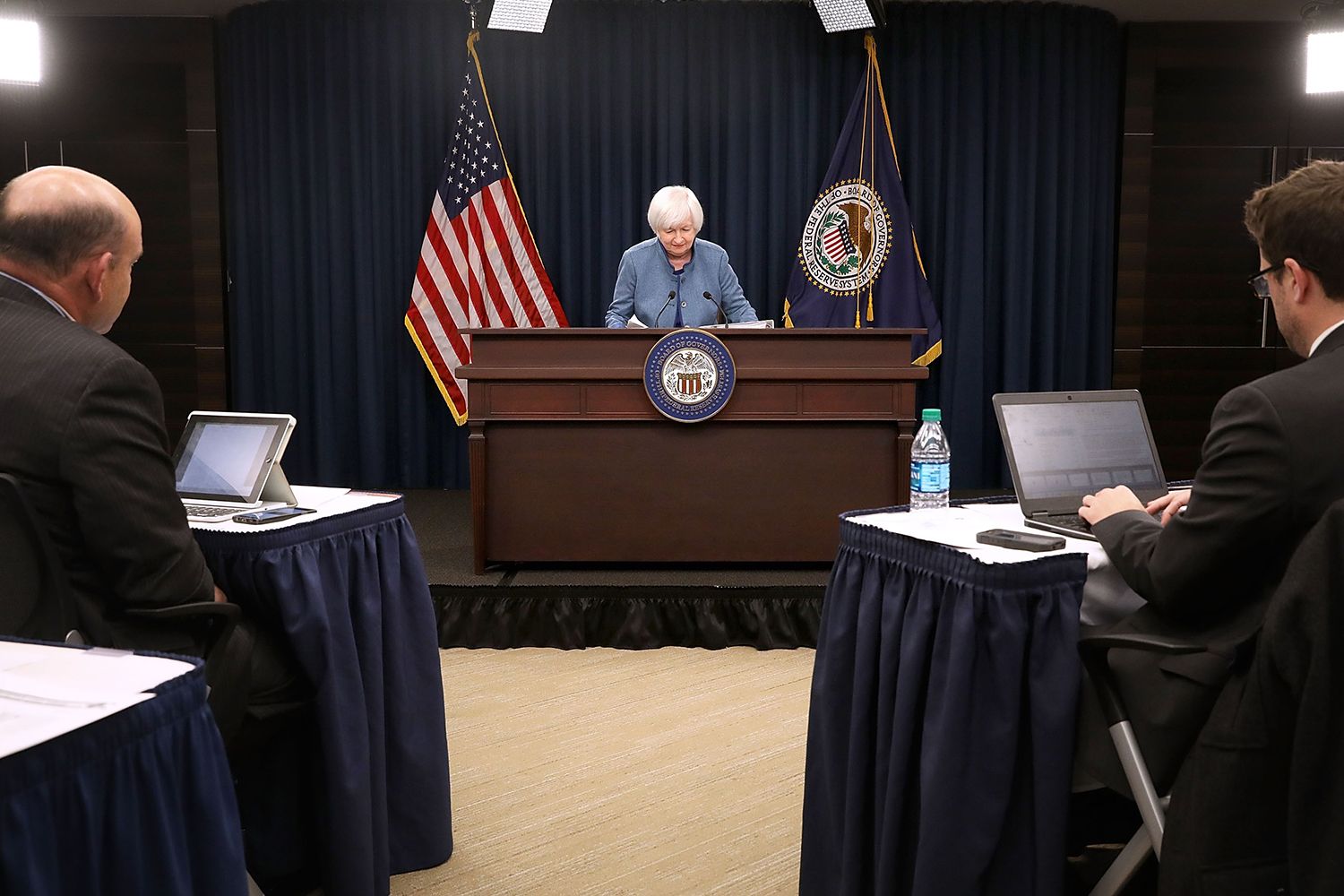 Federal Reserve Board Chair Janet Yellen stands at podium between the American flag and the flag of Washington DC reading from an announcement.
