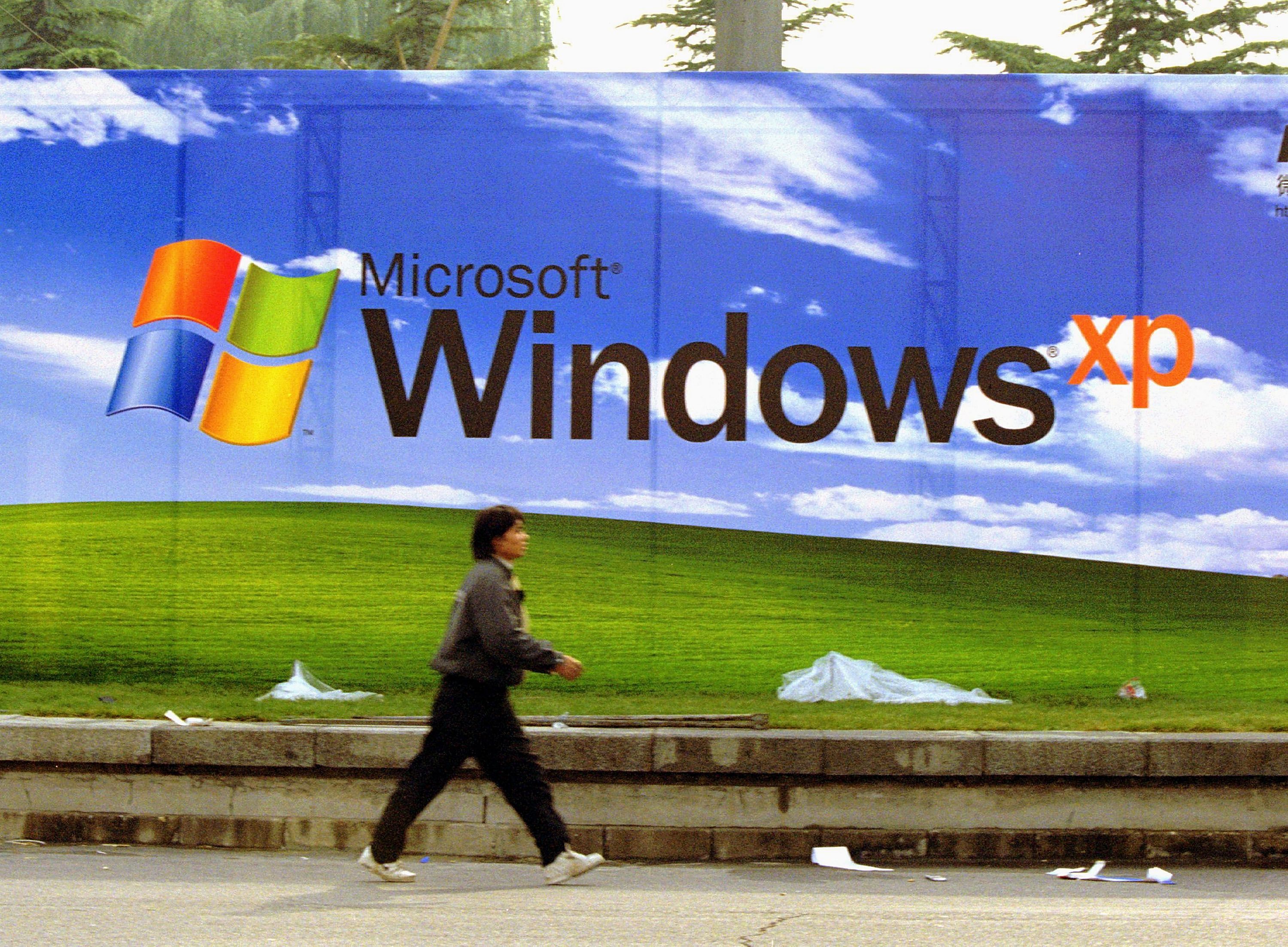 A person walks by an outdoor advertisement for Windows XP showing the green field and vibrant blue sky scattered with clouds users have come to associate with the Microsoft brand.