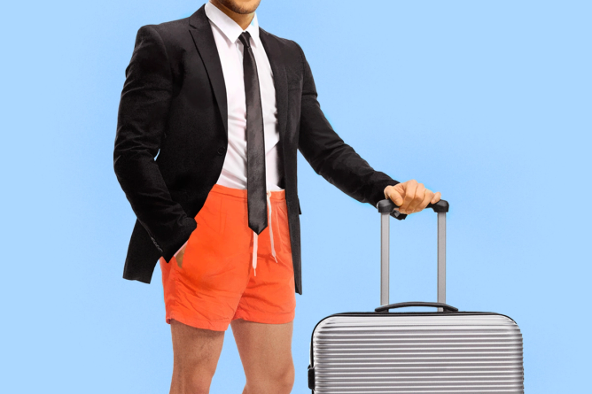 A man dressed in a suit and casual shorts holds onto a rolling suitcase.