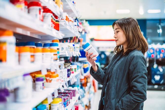 Asian woman reading the ingredient label while shopping for multivitamin and health supplements on shelf in supermarket.