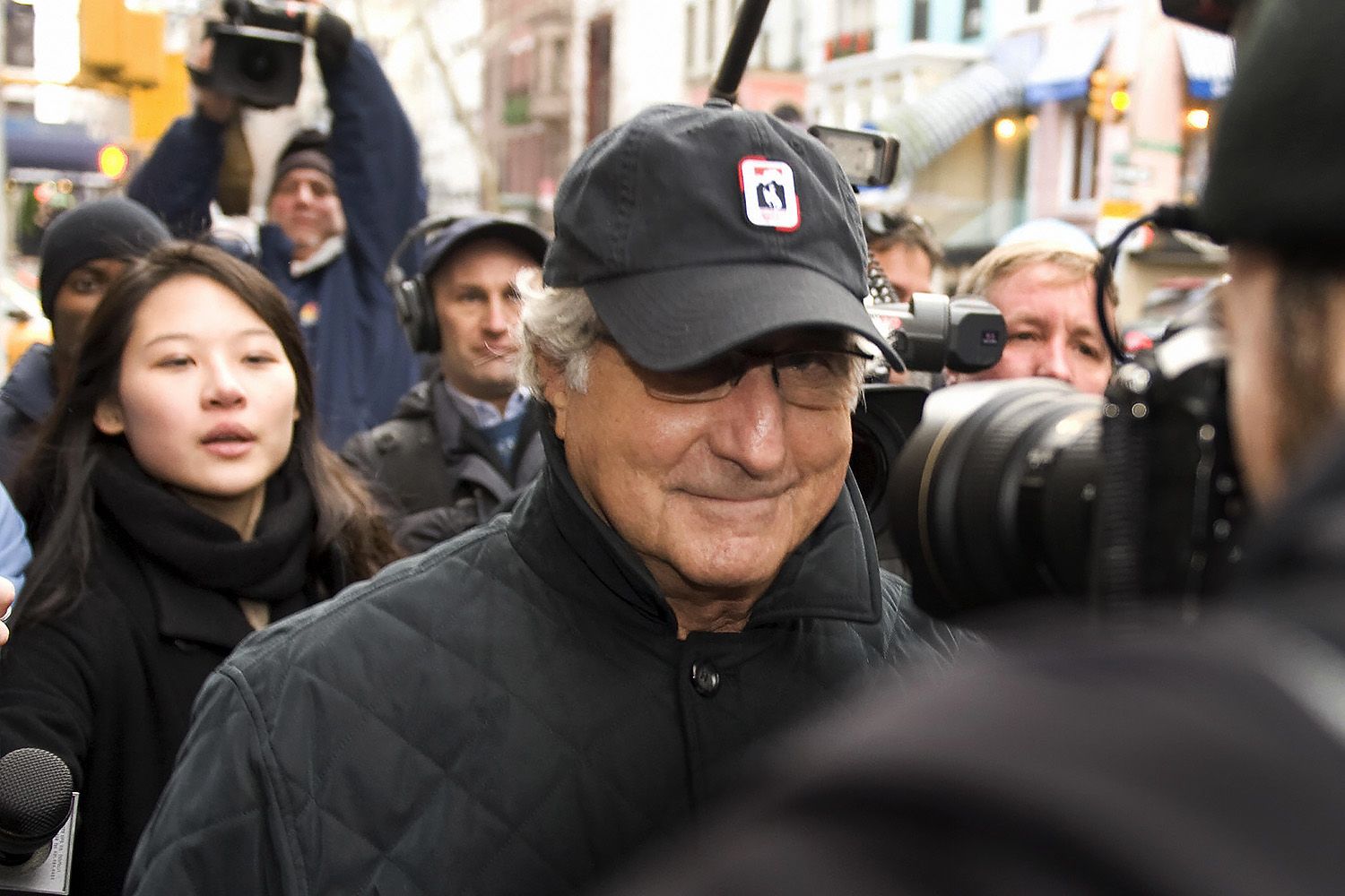 Bernie Madoff in a casual baseball cap smirks at photographers, surrounded by press.
