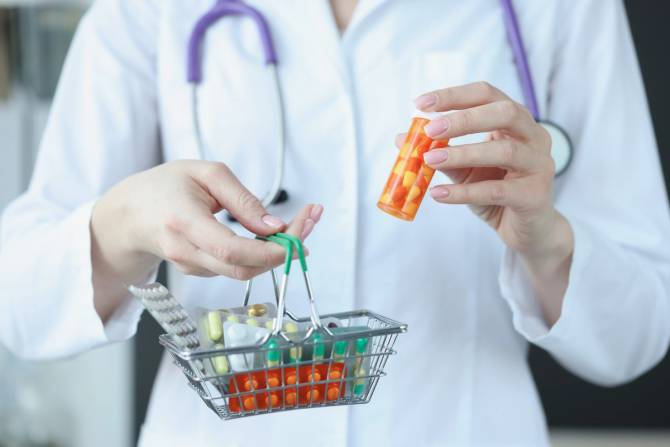 A doctor holds a pill case in one hand and a small basket with pills of different sizes in the other.
