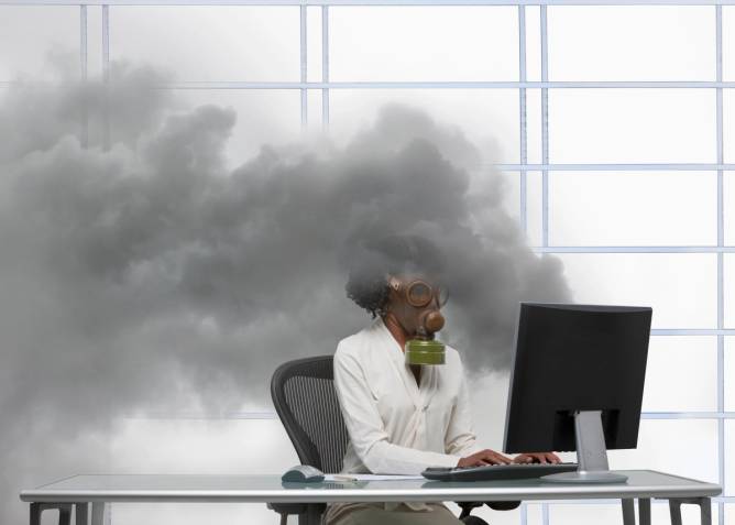 black female businessperson sitting at a desk with a gas mask on as a flume of smoke comes up behind her.