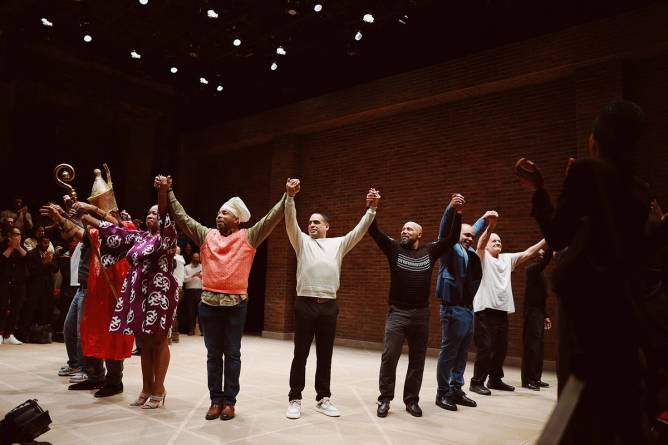 A group of men and one woman hold hands on stage while taking a bow at the end of a performance. 