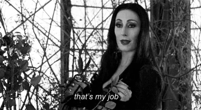 Angelica Huston, as Morticia Addams, standing in front of vine-covered windowpanes, with the caption "that's my job" underneath her