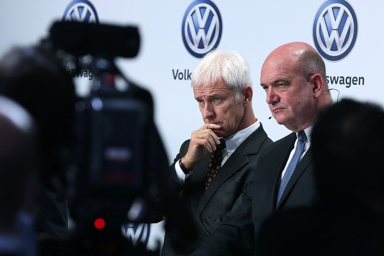 German carmaker Volkswagen leadership stand in front of a wall with the company logo while a video camera points at them.