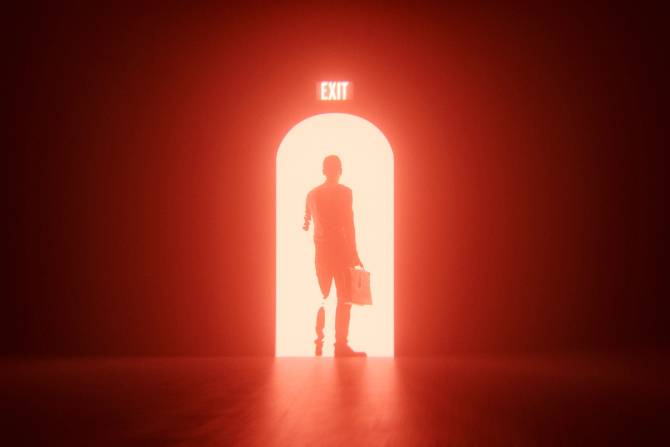 red exit sign above a door with a bright light shining on a shadowy businessperson walking through